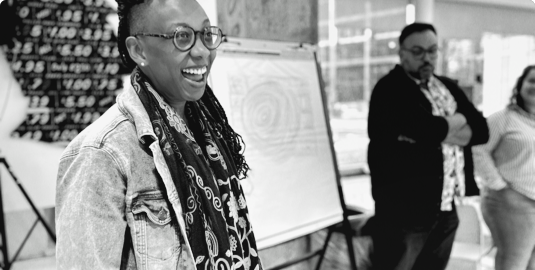 A Black woman wearing glasses smiles with a Latino man standing beside her with his arms crossed during a group discussion.